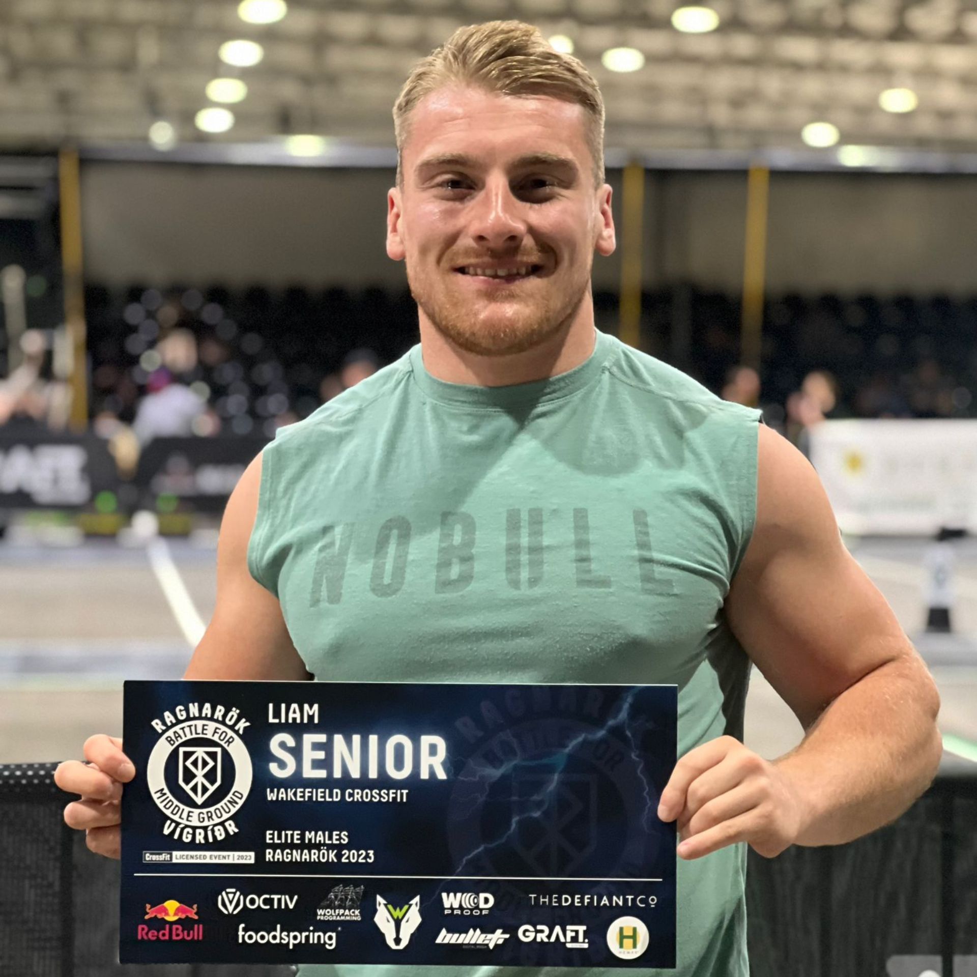 Man in a green tank top holds a sign with event details and sponsors in a sports setting.
