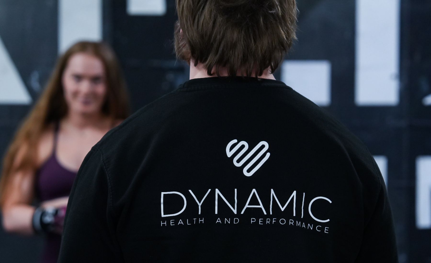 Person wearing a jacket labeled "Dynamic Health and Performance," facing someone in a gym setting.
