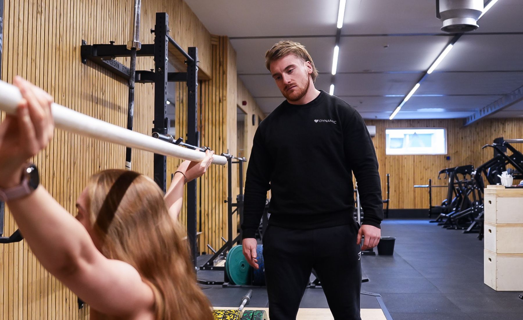 A woman performs a bench press with a barbell while a man supervises in a gym.
