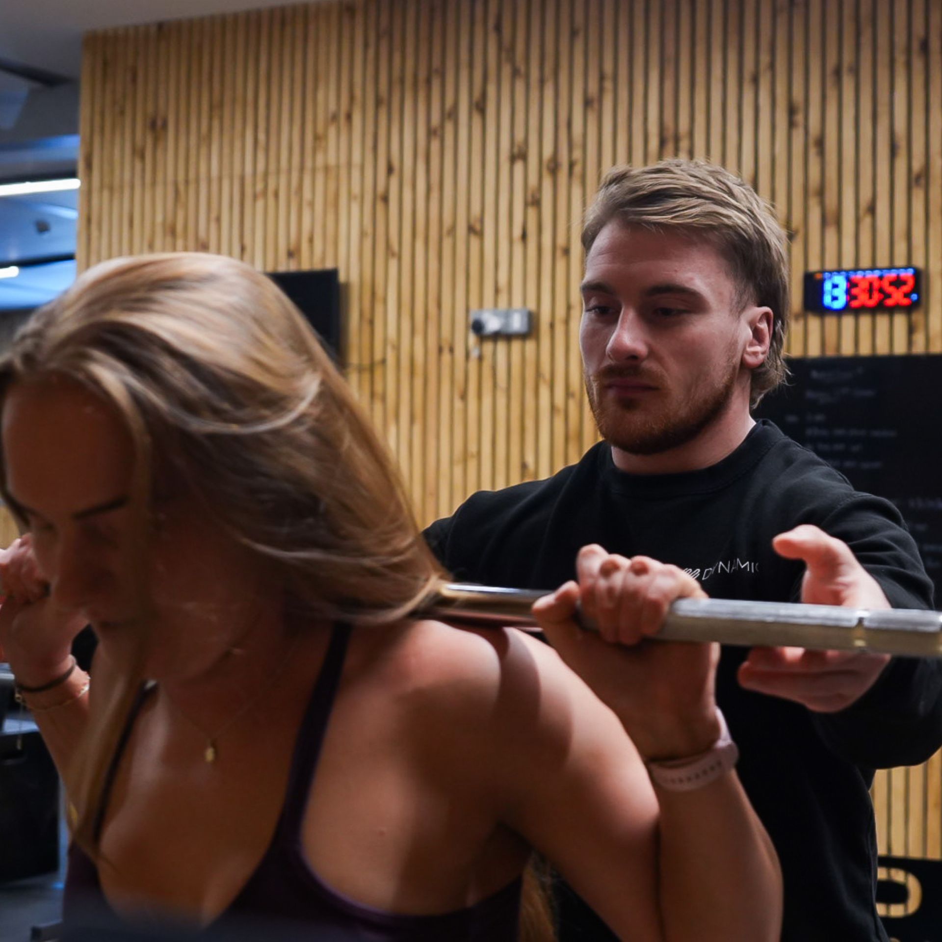 A woman is lifting a barbell while a trainer assists her in a gym setting.