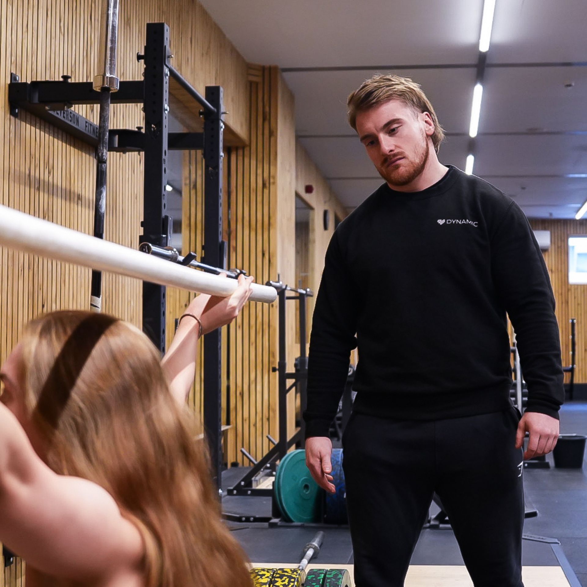 A woman lifts a barbell in a gym while a man watches and coaches her.